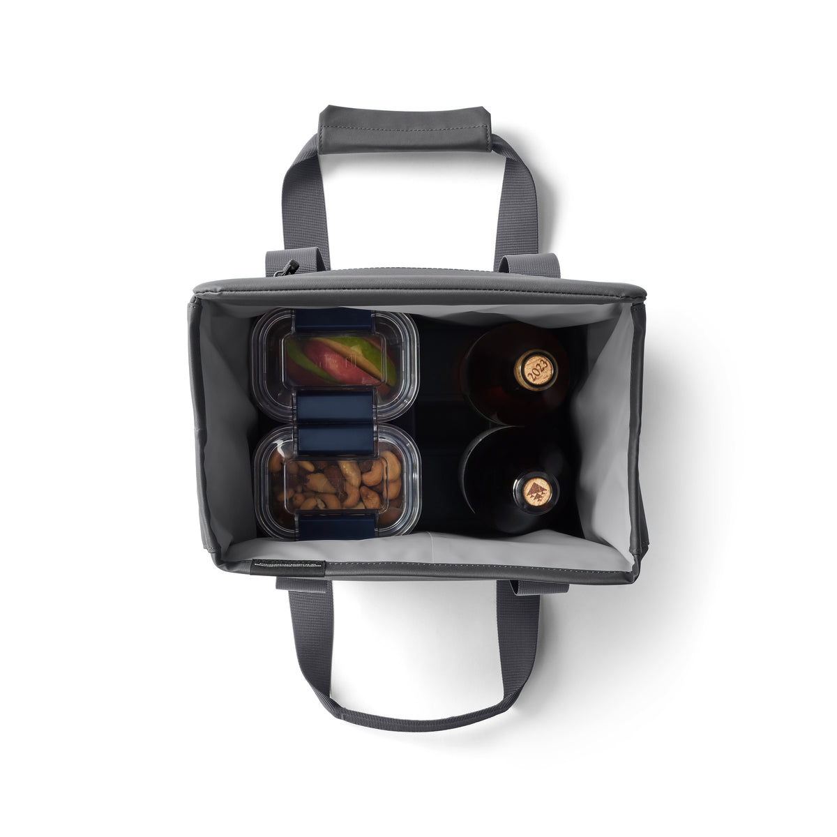Draufsicht auf eine geöffnete YETI® Kühltasche Daytrip Lunch Bag 14L - Charcoal mit zwei Flaschen, gemischtem Obst und Mandeln. Diese YETI Tasche hat Coldcell Flex Isolierung, sowie kurze Griffe und einen Schultergurt für einfaches Tragen.