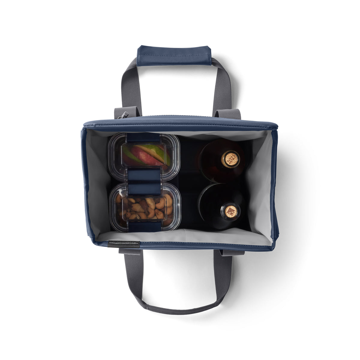 Draufsicht auf die YETI® Kühltasche Daytrip Lunch Bag 14L - Navy mit grauen Griffen, zeigt zwei Flaschen, einen durchsichtigen Behälter mit geschnittenem Obst und einen mit Mandeln. Die Coldcell Flex Isolierung von YETI hält den Inhalt frisch.