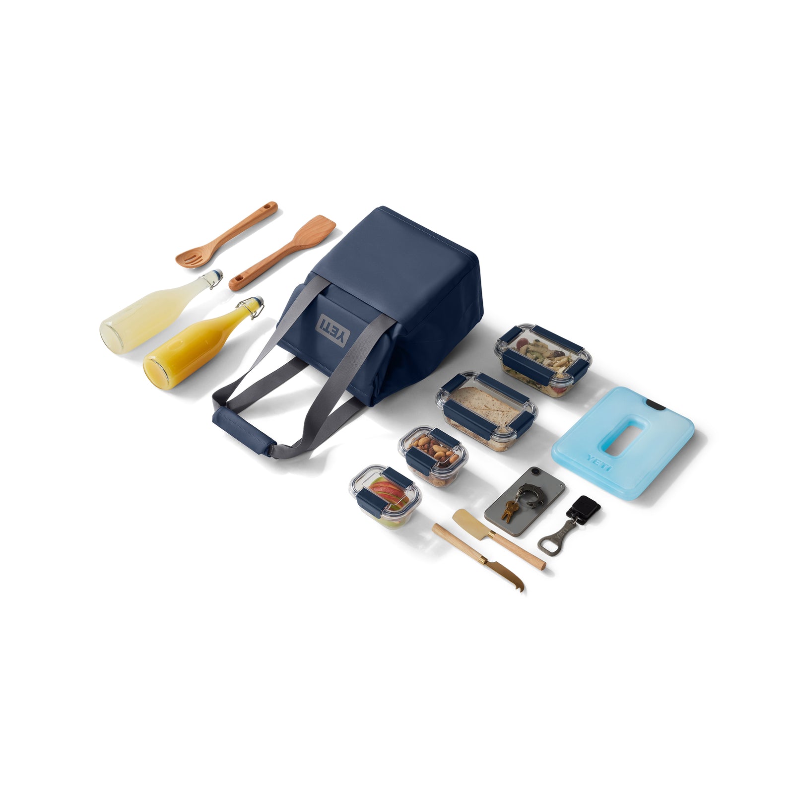 Eine YETI® Kühltasche Daytrip Lunch Bag 14L in Navy steht auf einem weißen Hintergrund, umgeben von Holzutensilien, zwei Glasflaschen, einem blauen Schneidebrett, wiederverwendbarem Besteck und Behältern, die mit Sandwiches und Snacks gefüllt sind.