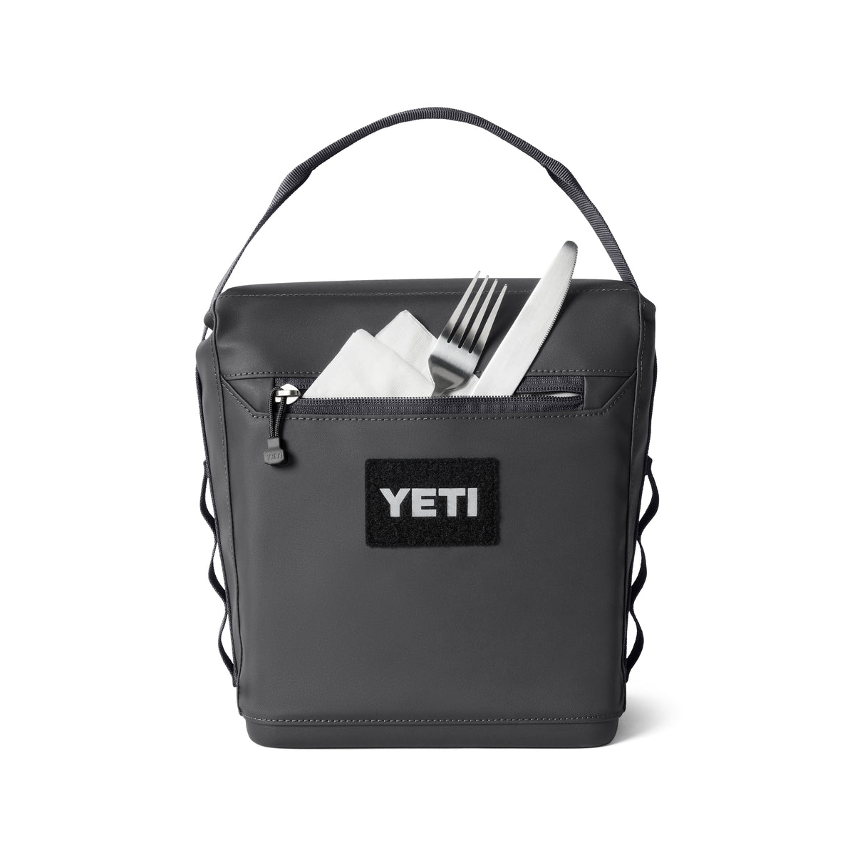 Die YETI® Kühltasche Daytrip Lunch Bag 6L in Charcoal verfügt über einen Tragegriff und eine Reißverschlusstasche auf der Vorderseite. Die Abbildung zeigt eine Serviette mit Gabel und Messer in der teilweise geöffneten Tasche.
