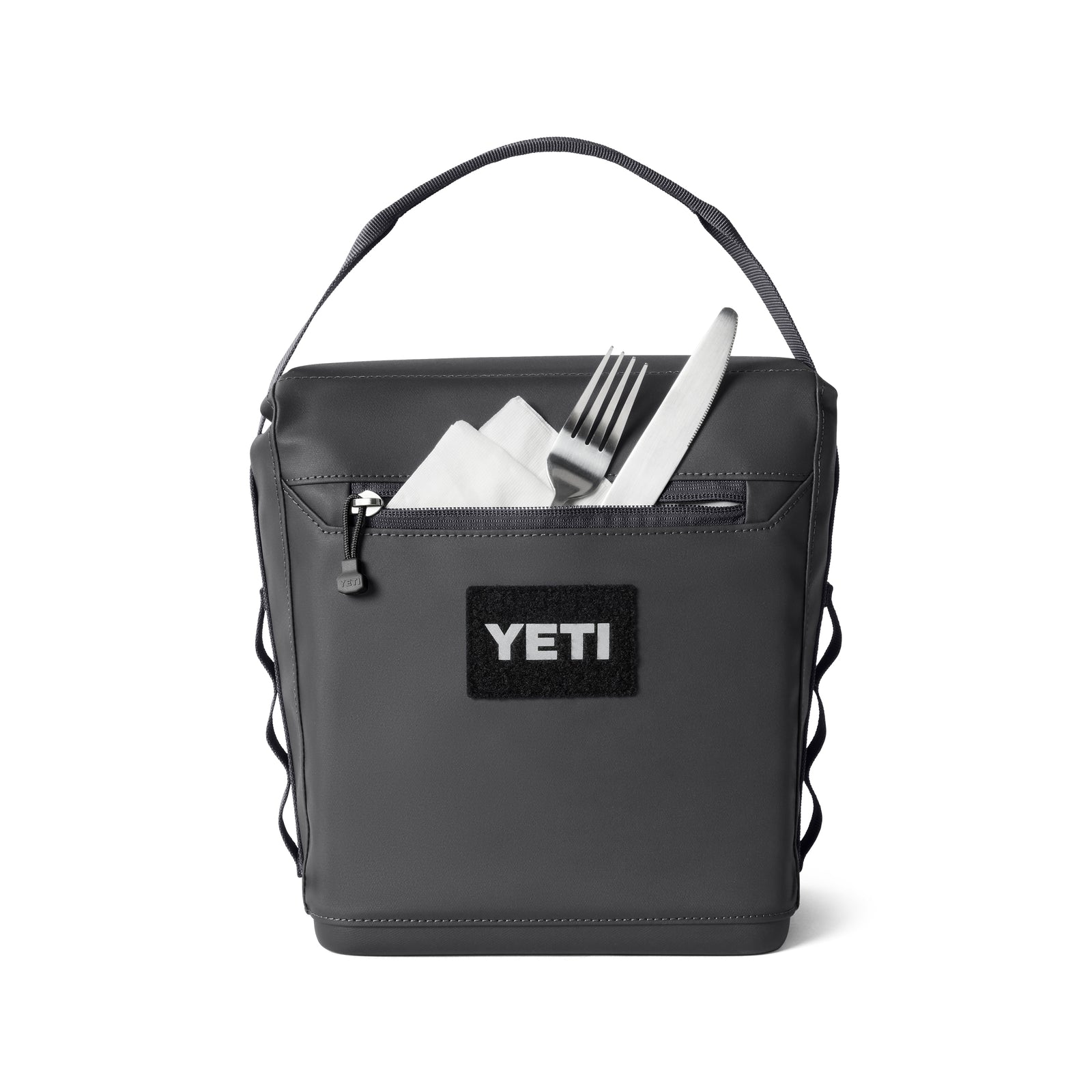 Die YETI® Kühltasche Daytrip Lunch Bag 6L in Charcoal verfügt über einen Tragegriff und eine Reißverschlusstasche auf der Vorderseite. Die Abbildung zeigt eine Serviette mit Gabel und Messer in der teilweise geöffneten Tasche.