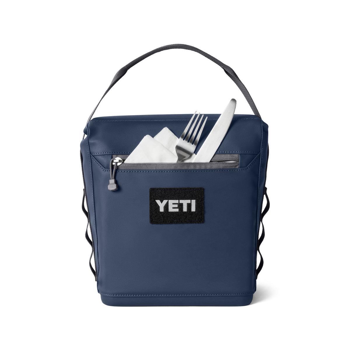 Die YETI® Kühltasche Daytrip Lunch Bag 6L in Navy verfügt über einen Tragegriff, eine Vordertasche mit Reißverschluss und schwarzem YETI-Aufnäher sowie eine Kühlisolierung. Eine Gabel, ein Messer und eine Serviette schauen aus der Tasche dieser YETI Kühltasche hervor.