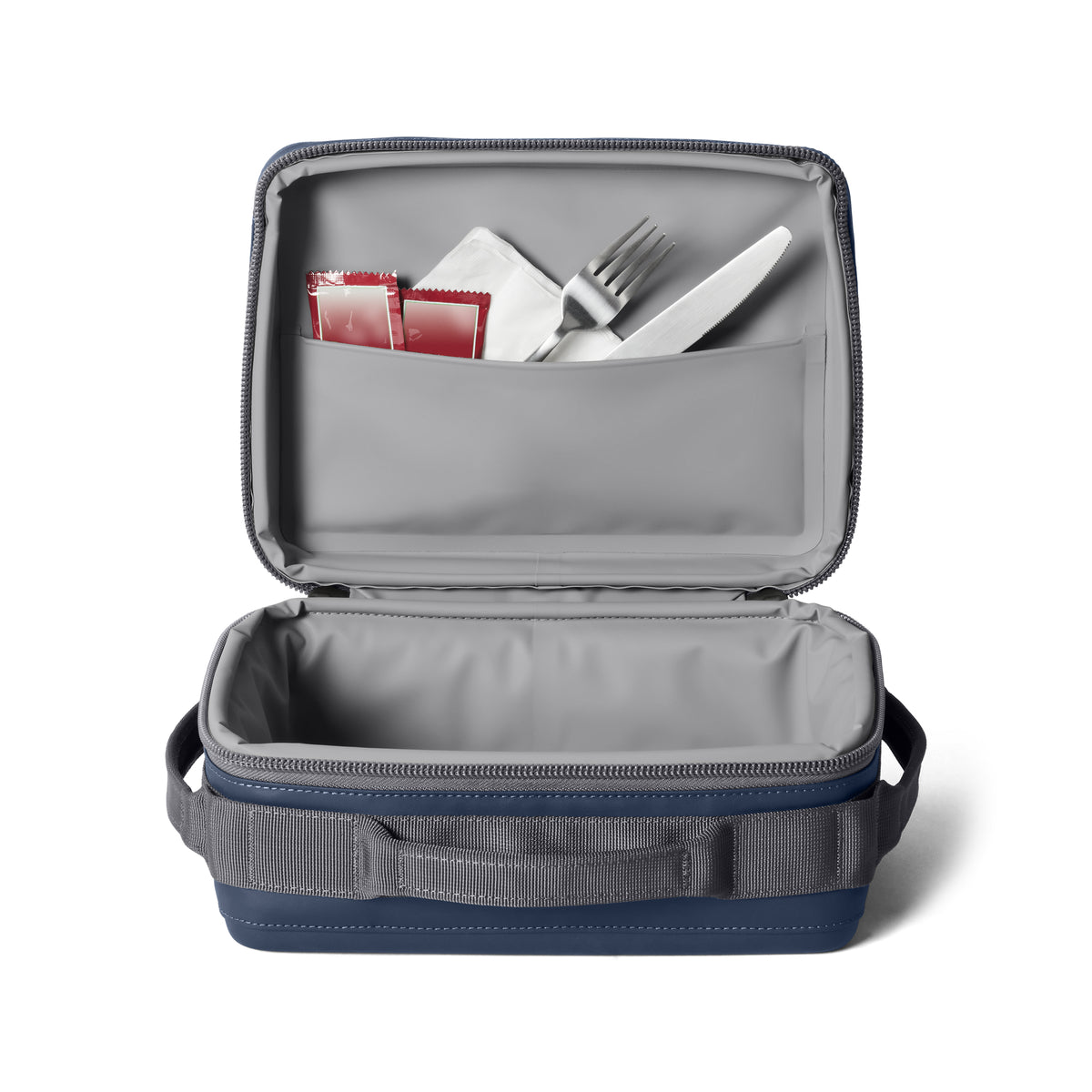 In der geöffneten navyfarbenen YETI® Kühltasche Daytrip Lunch Box 5L befinden sich in der Innentasche eine Gabel, ein Messer, eine Serviette und zwei Soßenpakete. Die Lunchbox hat kurze Seitengriffe zum einfachen Tragen und einen Deckel mit Reißverschluss.