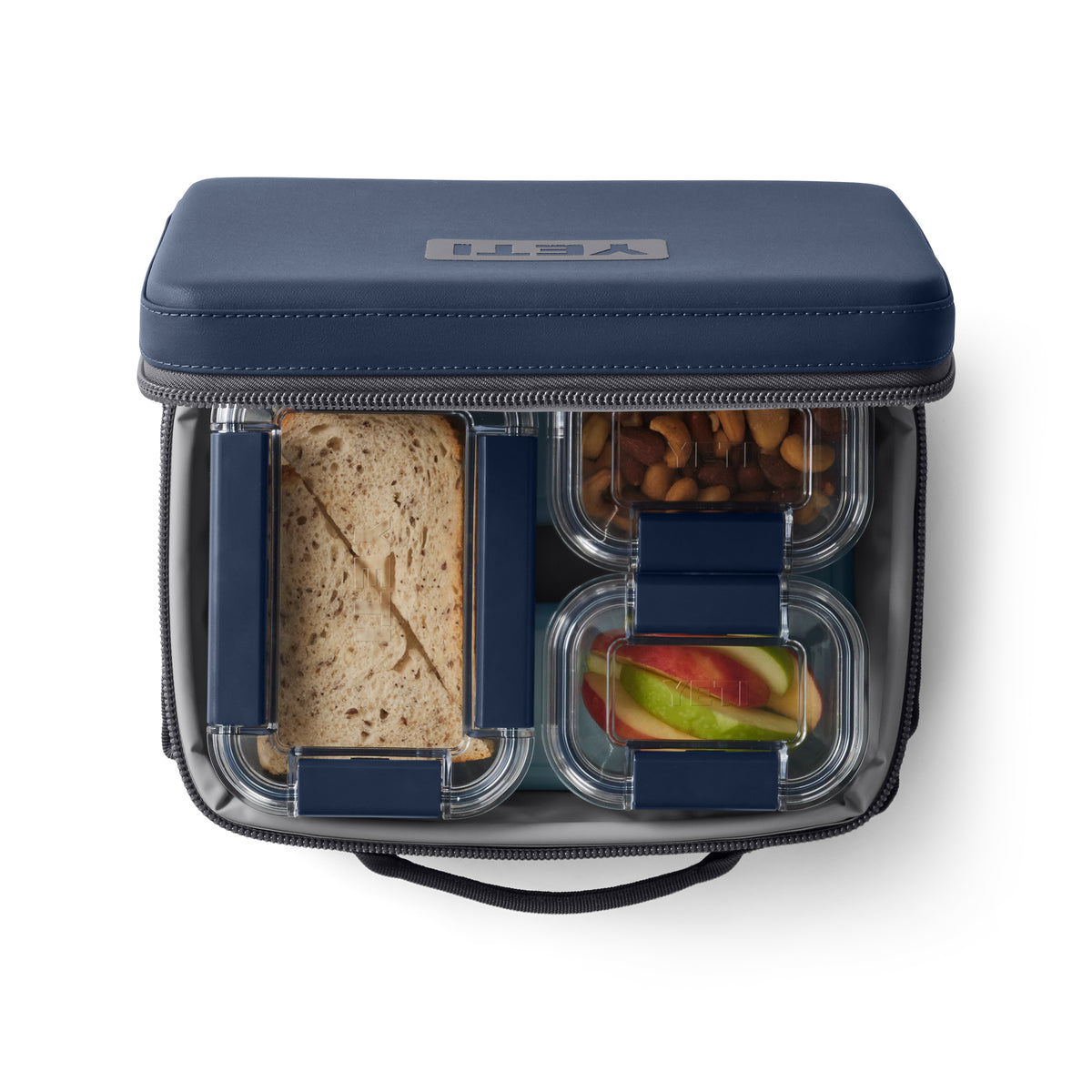 Die navyfarbene YETI® Kühltasche Daytrip Lunch Box 5L ist offen und gibt den Blick frei auf ein Sandwich, geschnittene Äpfel und Nüsse, die in durchsichtigen Behältern in dieser praktischen Lunchbox von YETI aufbewahrt werden.