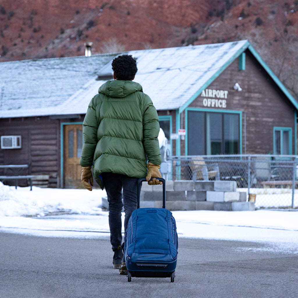 Eine Person in einem grünen Wintermantel zieht einen schwarzen YETI® Koffer Crossroads® 22 in Richtung eines kleinen Holzgebäudes mit der Aufschrift Airport Office an einem verschneiten Tag, mit Bergen im Hintergrund.
