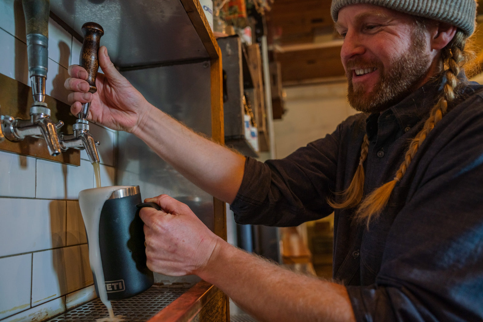 Eine lächelnde Person mit Mütze gießt Bier aus einem Zapfhahn in einen YETI® Krug Rambler 24 Oz - Stainless Steel von YETI, so dass es in einer Bar schäumt und überläuft.