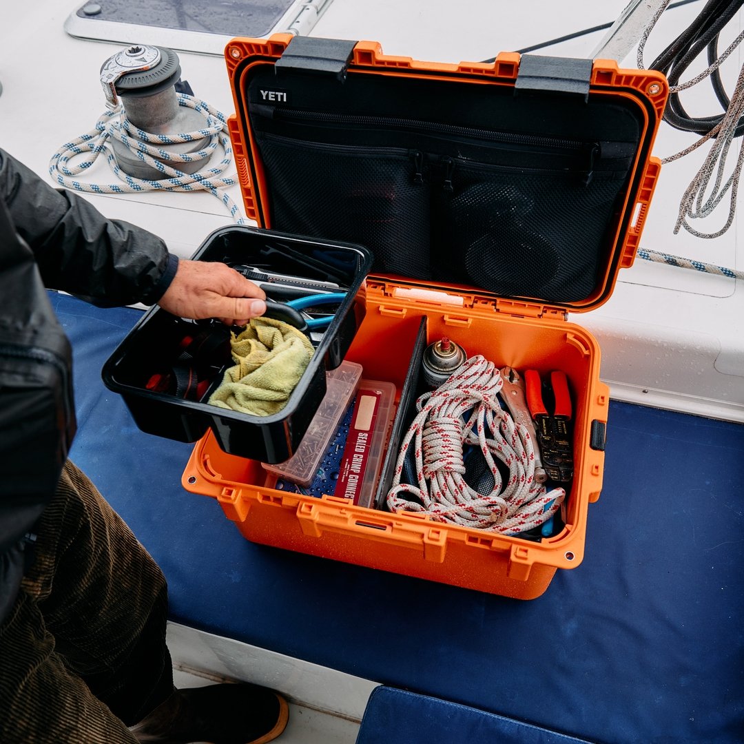 Ein Segler öffnet eine King Crab Orange YETI® LoadOut GoBox 30, die mit Seilen, Werkzeugen und Zubehör gefüllt ist. Der Deckel der Box ist mit Netztaschen versehen, während auf dem Deck des Segelboots dahinter Seile und eine Winde zu sehen sind.