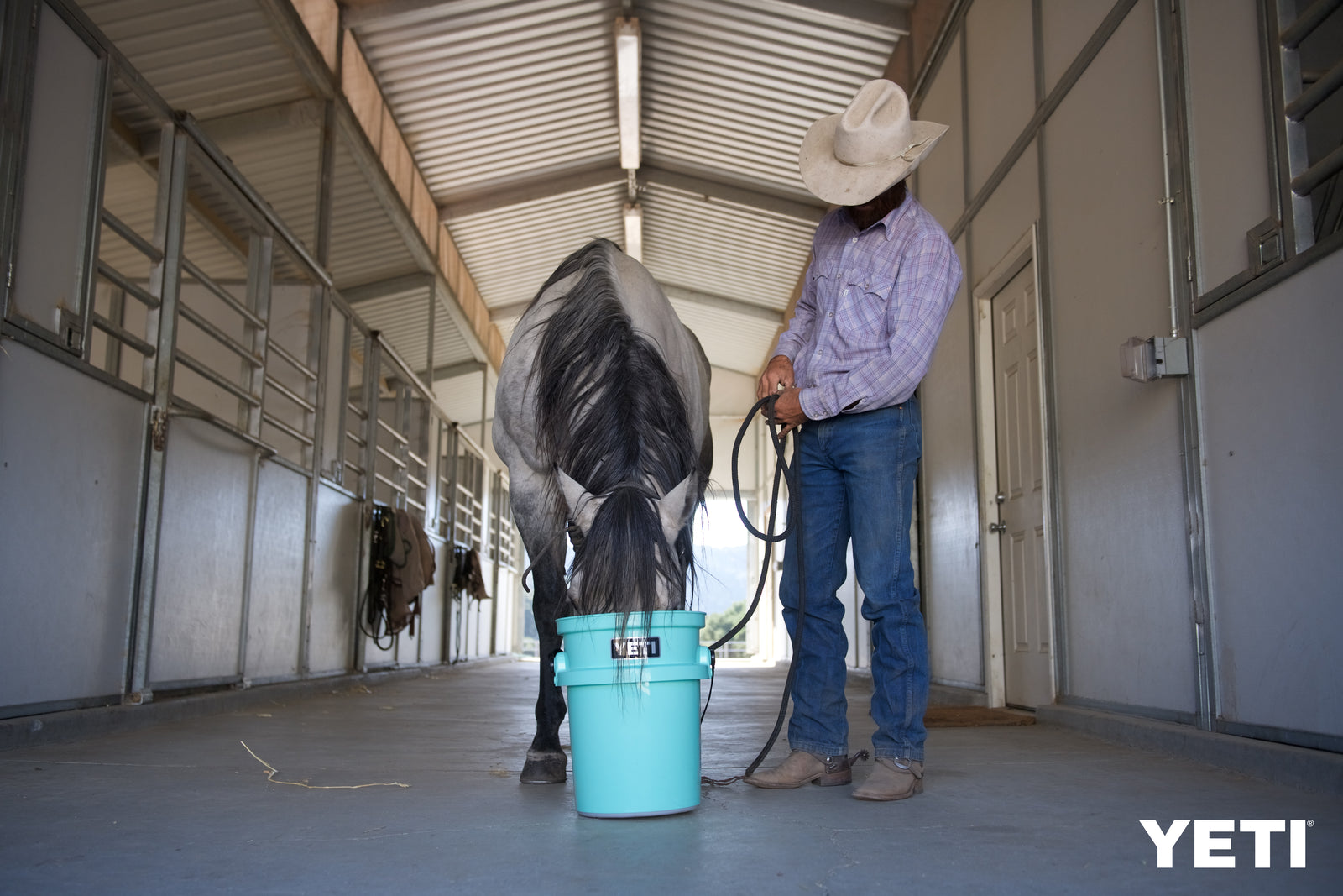 Ein Mann mit Cowboyhut steht neben einem Pferd, das aus einem meerschaumfarbenen YETI® Loadout Eimer 19 Liter in einem Stall mit Metallboxen isst. Das YETI-Logo ist in der unteren rechten Ecke zu sehen.