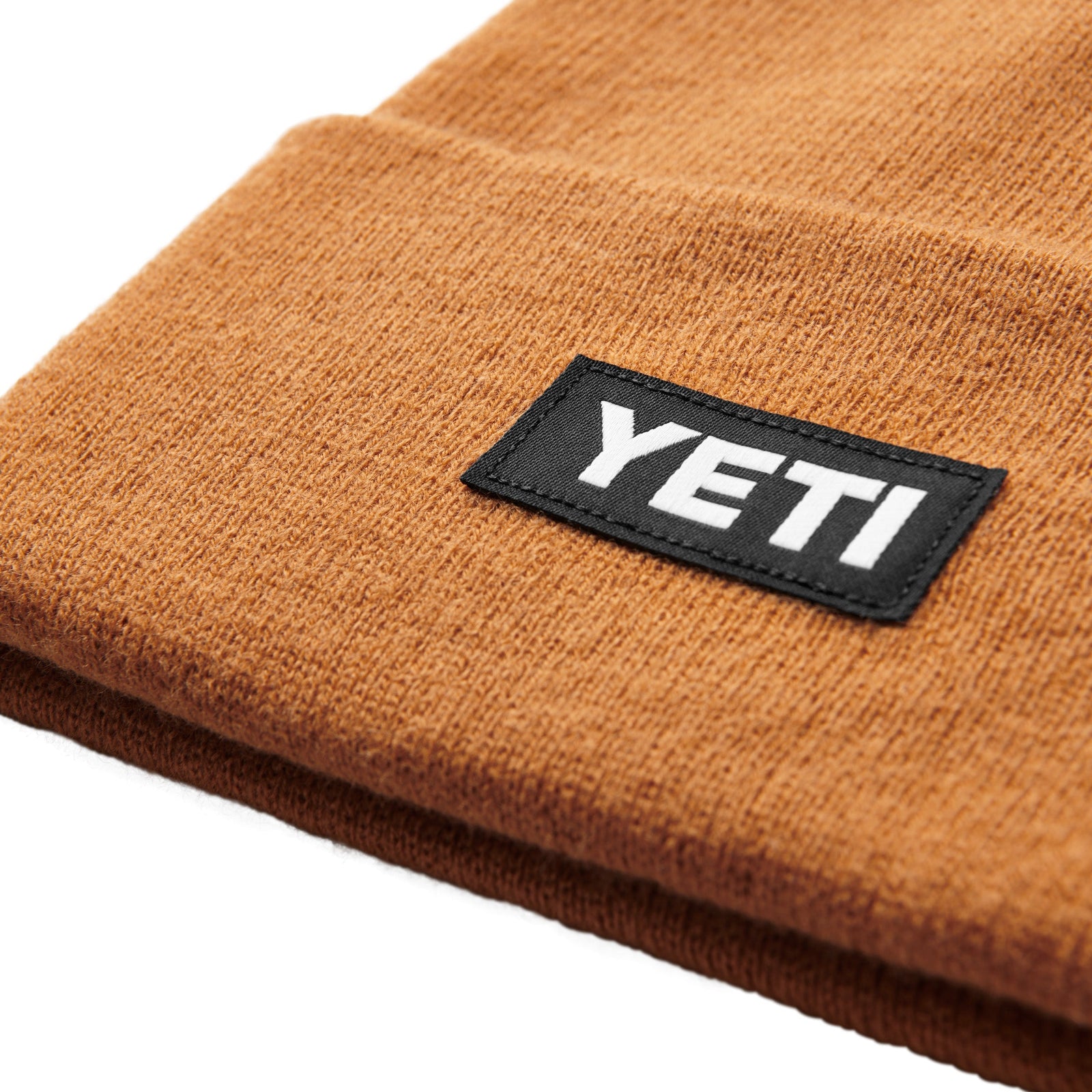 Nahaufnahme der YETI® Logo Badge Beanie, einer braunen Outdoor-Mütze, die mit einem schwarzen Aufnäher versehen ist, auf dem das weiße YETI-Branding prangt.