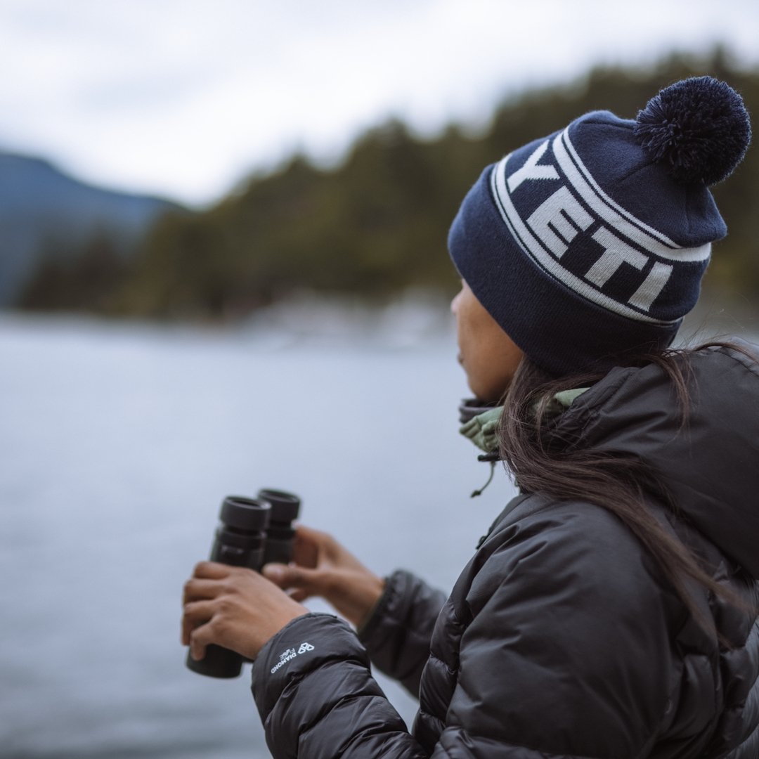 Eine Person, die eine Winterjacke und eine YETI® Logo Retro Knit Beanie trägt, hält ein Fernglas in der Hand und blickt auf einen See, mit verschwommenen Bäumen und Bergen im Hintergrund.