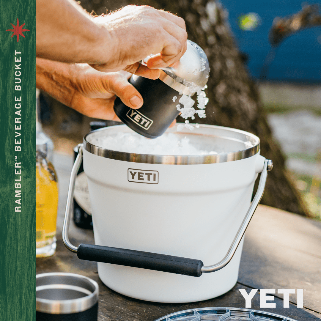 Eine Person gießt Eis aus einem YETI Becher in einen YETI® Rambler® 7,6 Liter Getränkekübel - Lowcountry Peach mit doppelwandiger Isolierung im Freien, im Hintergrund sind Getränke und Flaschen zu sehen. Text: RAMBLER BEVERAGE BUCKET und YETI.