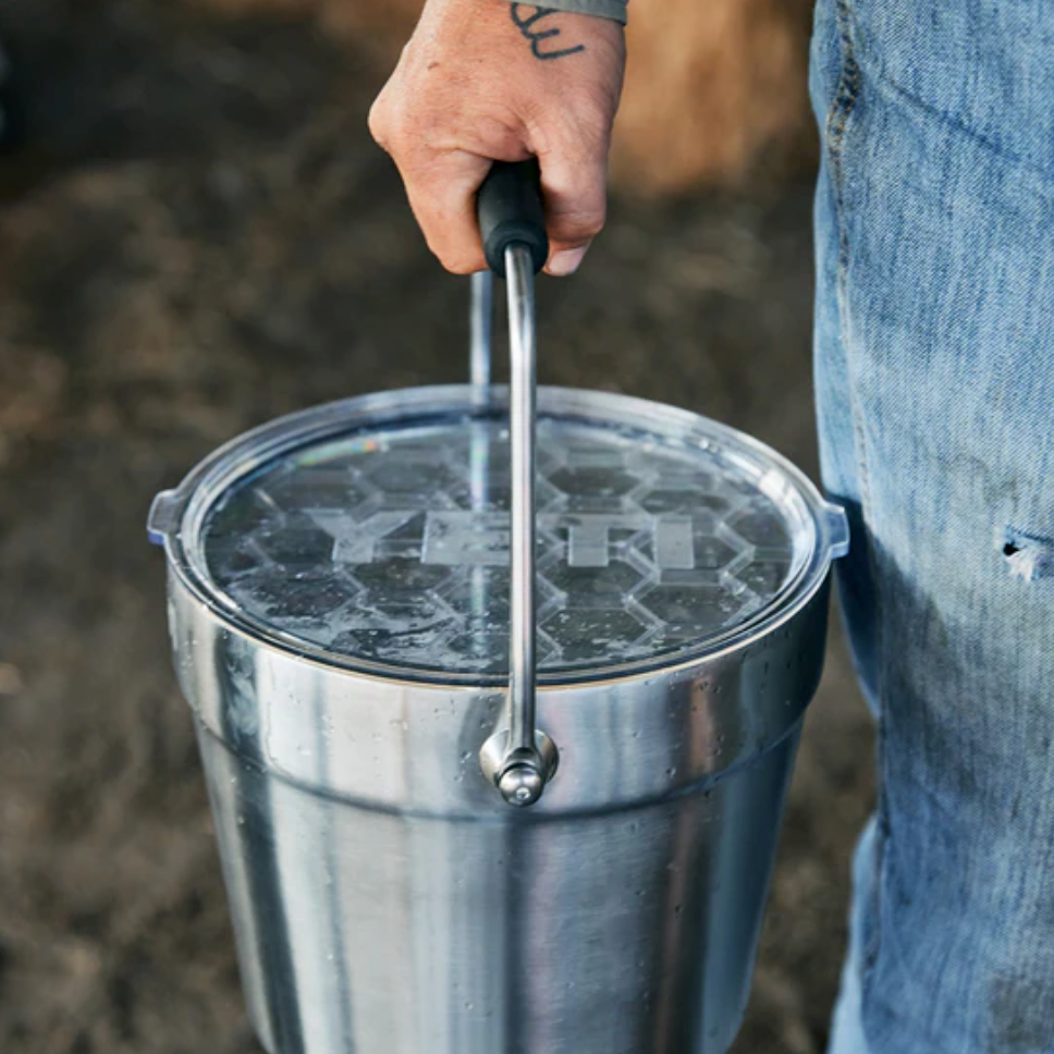 Eine tätowierte Hand hält den Griff eines YETI® Rambler® 7,6-Liter-Getränkekübels in Tropical Pink mit klarem Deckel, der von einer Person in Jeans im Freien gehalten wird.