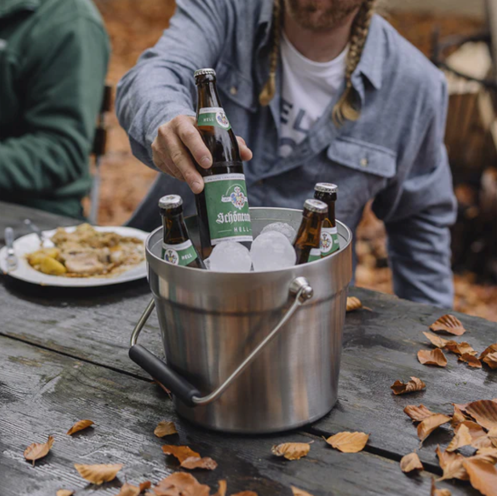 Jemand nimmt ein Bier aus dem YETI® Rambler® 7,6 Liter Getränkekübel - White, der über eine doppelwandige Vakuumisolierung verfügt, auf einem verwitterten Picknicktisch inmitten von Herbstblättern und Essen im Freien.