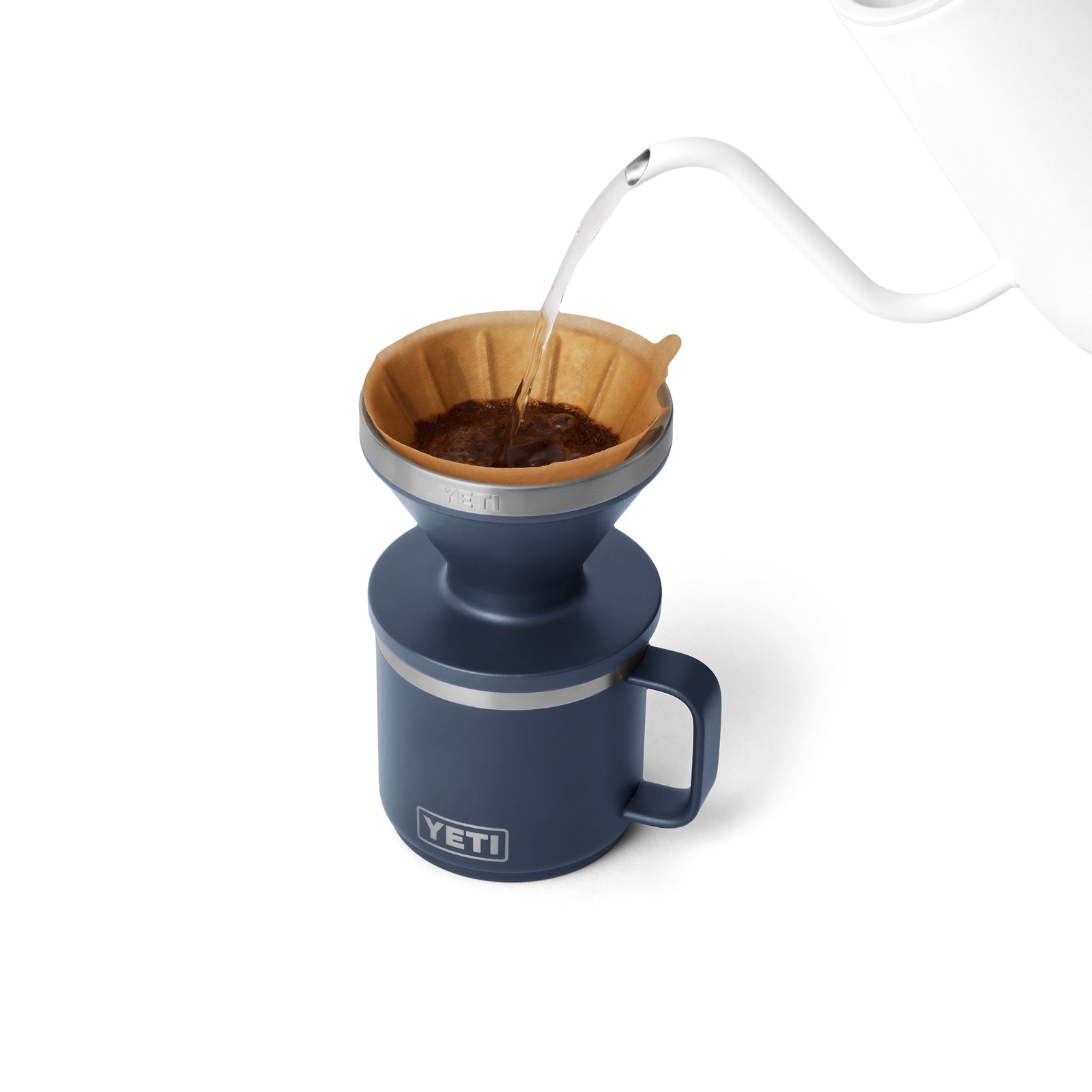 Ein weißer Wasserkocher gießt heißes Wasser über den Kaffee in einem Kaffeebereiter-Filter auf dem navyfarbenen YETI® Rambler Pour Over-Kaffeebereiter und brüht einen Pour Over-Kaffee vor einem weißen Hintergrund.