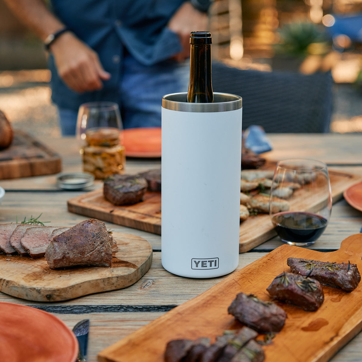Der weiße YETI® Rambler® Weinkühler von YETI hält eine Flasche auf einem Outdoor-Tisch mit Grillfleisch, Steak, Holzplatten, Rotweingläsern und einer Person im Hintergrund zuverlässig kühl.