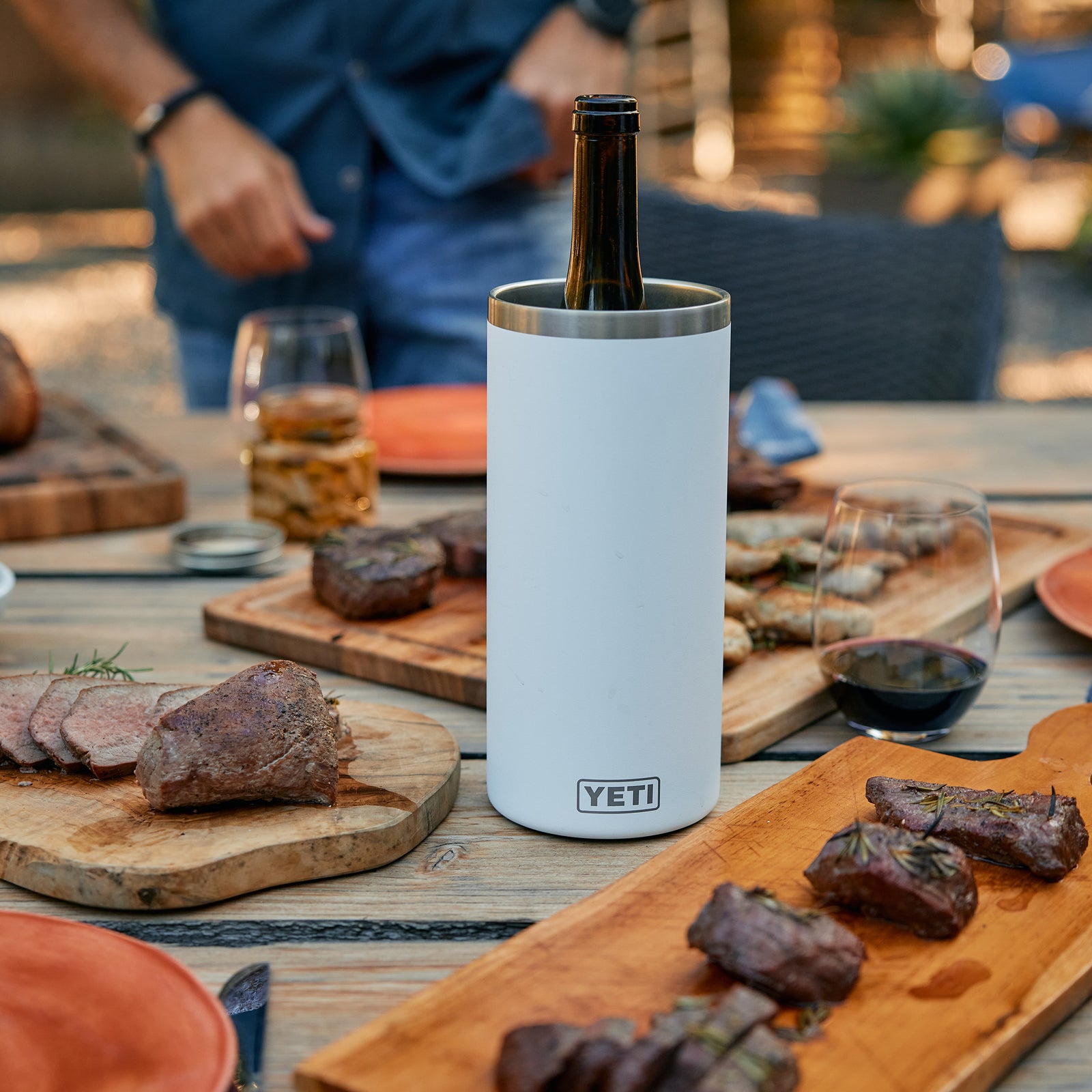 Der weiße YETI® Rambler® Weinkühler von YETI hält eine Flasche auf einem Outdoor-Tisch mit Grillfleisch, Steak, Holzplatten, Rotweingläsern und einer Person im Hintergrund zuverlässig kühl.