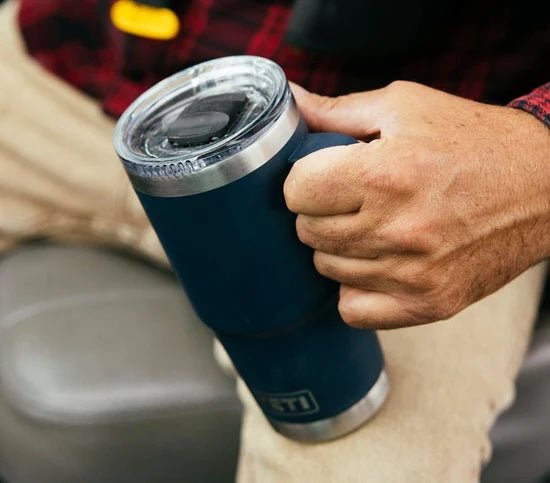 Eine Person in beigen Hosen und einem rot karierten Hemd hält einen YETI® Reisebecher Rambler 20 Oz - Lowcountry Peach und sitzt auf einem grauen Stuhl.