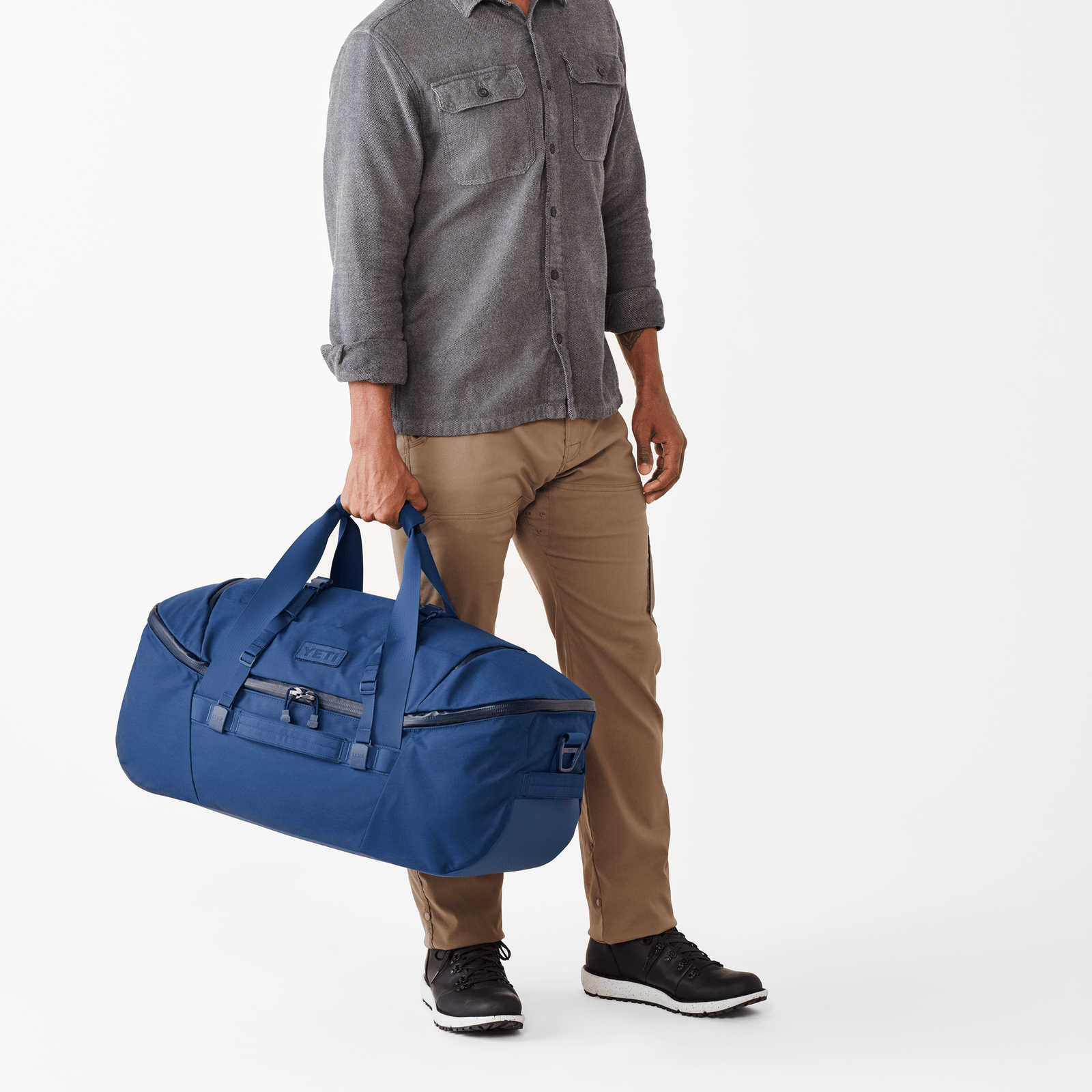 Mann in grauem Hemd und khakifarbener Hose trägt eine große olivgrüne YETI® Reisetasche Crossroads® 60L aus robustem, wasser- und abriebfestem TuffSkin Nylon an den Griffen vor einem weißen Hintergrund.