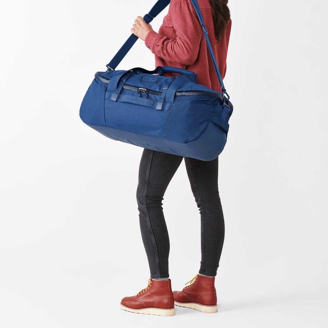 Eine Person in rotem Langarmshirt, schwarzer Jeans und roten Stiefeln trägt die YETI® Reisetasche Crossroads® 60L - Olive aus robustem, wasser- und abriebfestem TuffSkin Nylon über der Schulter vor weißem Hintergrund.