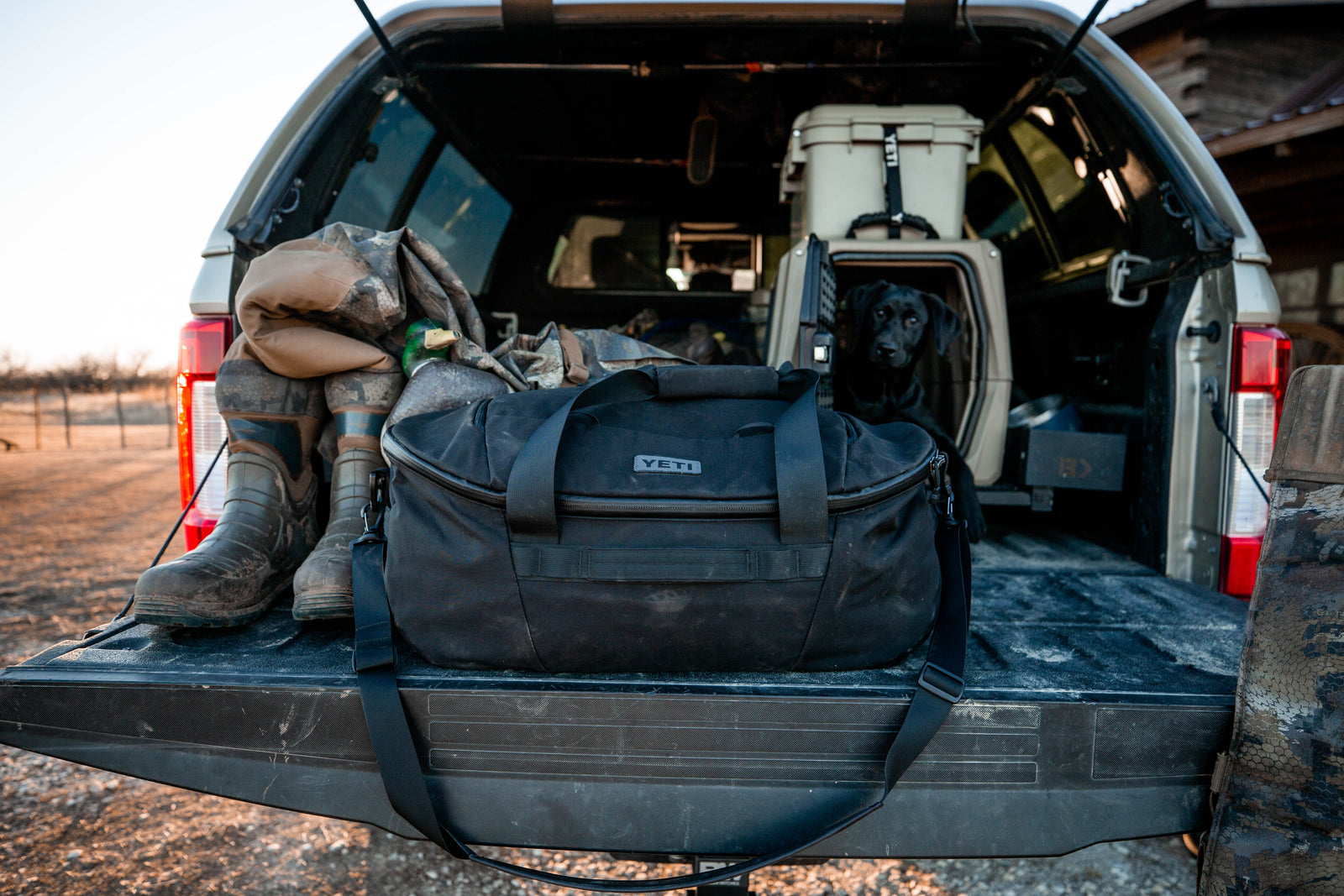 Auf der Heckklappe eines Pickups ist eine Jagdausrüstung zu sehen - Stiefel, Tarnkleidung, eine schwarze YETI® Reisetasche Crossroads 60 L und ein schwarzer Hund in einer Kiste - was auf ein Outdoor-Abenteuer oder einen Jagdausflug hindeutet.