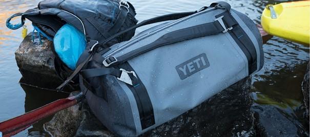 Eine YETI® Reisetasche Panga 50 in Storm Grey liegt auf einem Felsen am Wasser neben einer schwarzen Tasche, einem blauen Gegenstand und einem Teil eines gelben Paddleboards - perfekt für Outdoor-Abenteuer.
