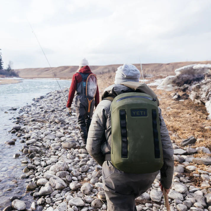Zwei Personen in Winterkleidung, die jeweils einen YETI® Rucksack Cayo™ 25L - Olive tragen, gehen mit Angelruten an einem felsigen Flussufer entlang. Die teilweise verschneite Landschaft umfasst kahle Bäume und Hügel im Hintergrund.