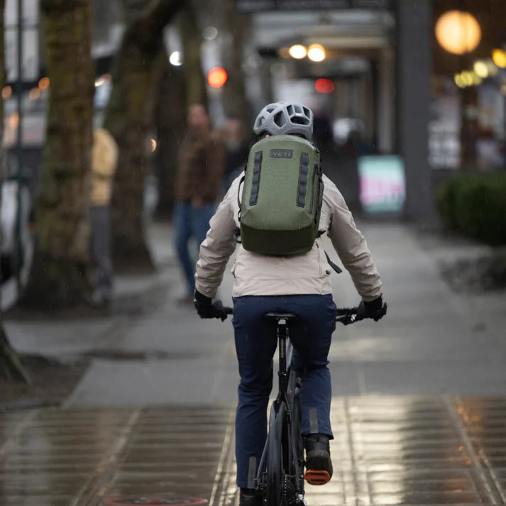 Eine Person, die einen Helm und einen YETI® Rucksack Cayo™ 25L - Olive trägt, fährt mit dem Fahrrad auf einem nassen Gehweg in der Stadt, mit verschwommenen Fußgängern und Straßenlaternen im Hintergrund.