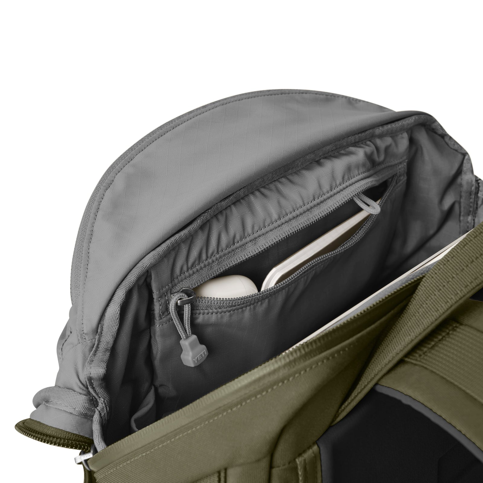 Ein offener olivfarbener YETI® Rucksack Crossroads® 22L von YETI zeigt seine Innentasche mit Reißverschluss, in der sich ein weißer Gegenstand, möglicherweise ein Notebook oder Tablet, vor einem schlichten weißen Hintergrund befindet.