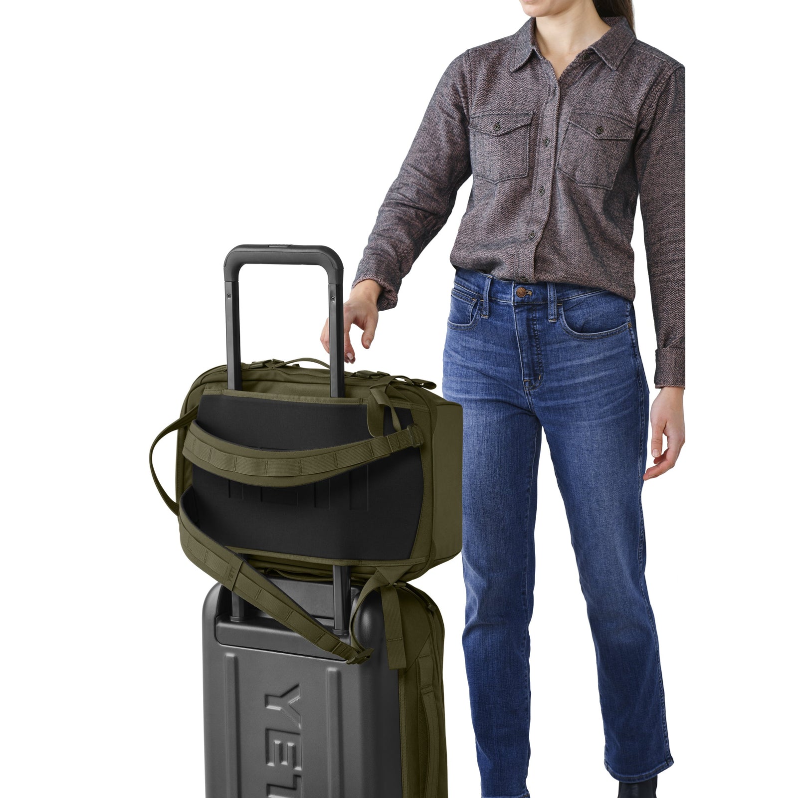 Eine Person in grauem Button-up-Hemd und blauer Jeans steht neben einem Koffer, auf dem ein YETI® Rucksack Crossroads® 22L in Olive steht, bereit für die Reise mit ihrem robusten Rucksack von YETI.