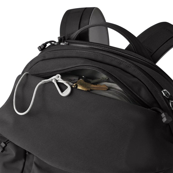 Eine Nahaufnahme des YETI® Rucksack Crossroads® 27L in Schwarz zeigt die Vordertasche mit offenem Reißverschluss und Schlüssel an einer weißen Kordel im Inneren; im Hintergrund sind die Schultergurte und das gepolsterte Laptopfach zu sehen.