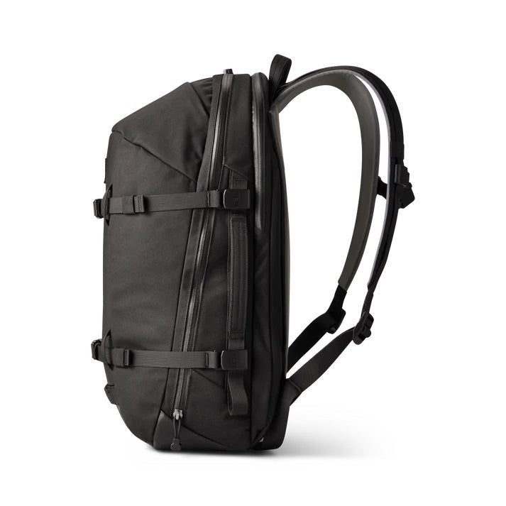 Seitenansicht des YETI® Rucksack Crossroads® 27L - Black mit gepolsterten Schultergurten, Kompressionsriemen, Reißverschlusstaschen, einem Laptopfach und einem strukturierten Design auf weißem Hintergrund.