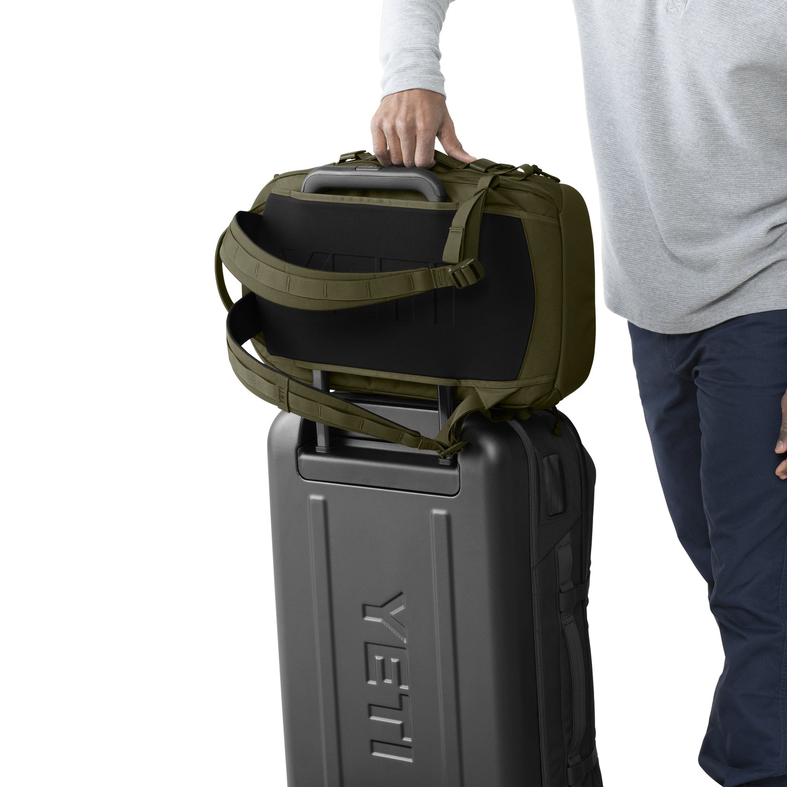 Eine Person mit hellgrauem Hemd und dunkelblauer Hose stellt den YETI® Rucksack Crossroads® 27L - Olive auf einen schwarzen YETI-Rollkoffer und hält den Koffergriff mit einer Hand.
