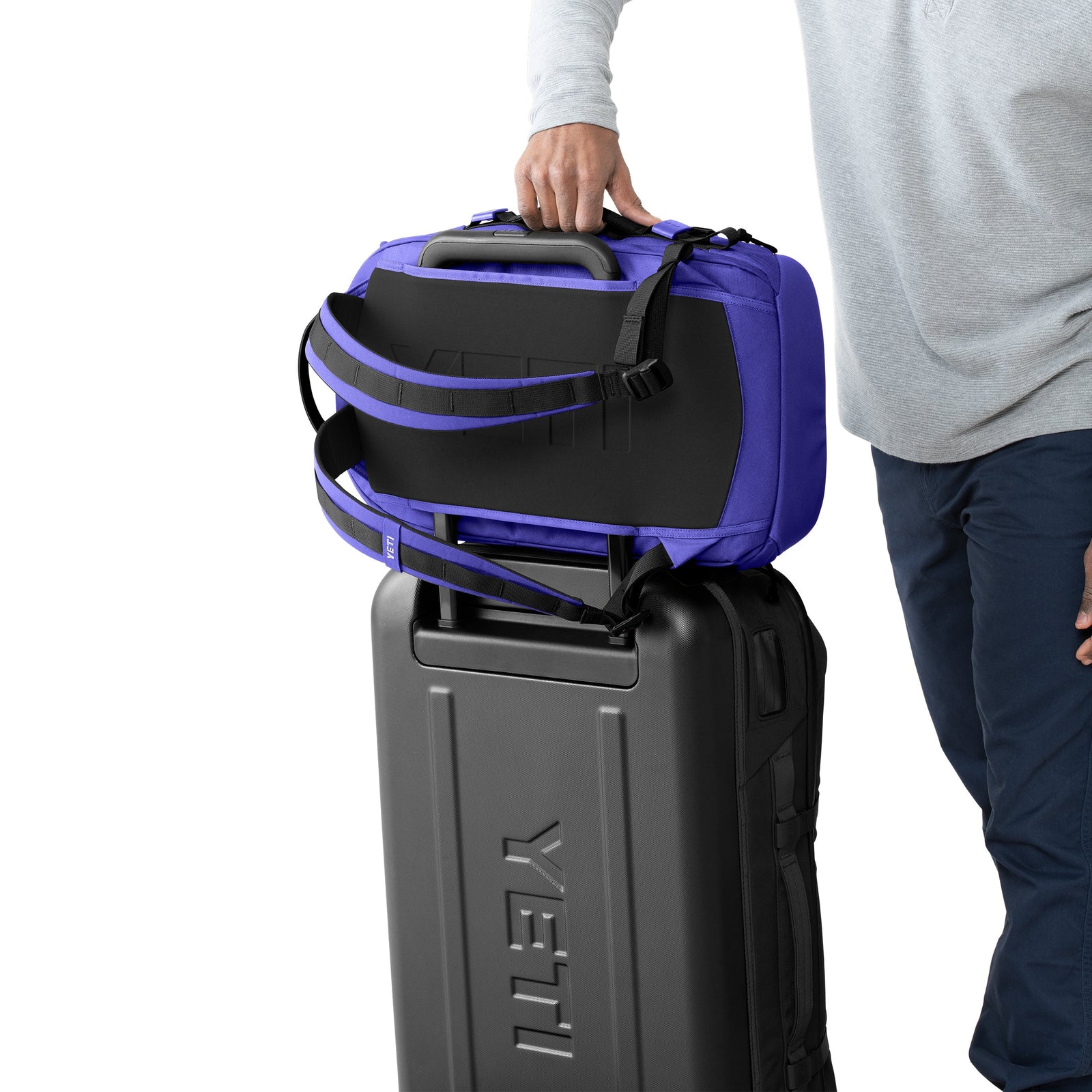 Eine Person in einem hellgrauen langärmeligen Hemd und einer blauen Hose stellt einen YETI® Rucksack Crossroads® 27L - Ultramarine mit Griffen auf einen schwarzen YETI Hartschalenkoffer.