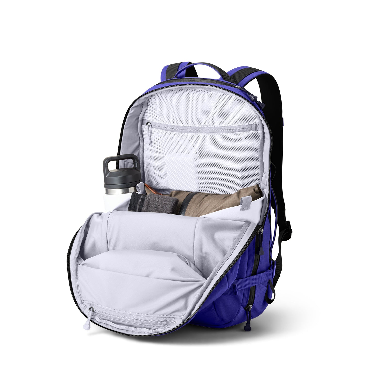 Der YETI® Crossroads® 27L Rucksack in Ultramarine ist geöffnet und zeigt eine Wasserflasche, Kleidung und Zubehör, die ordentlich in Netztaschen verstaut sind. Die leeren Bereiche betonen das geräumige Innere des Rucksacks.