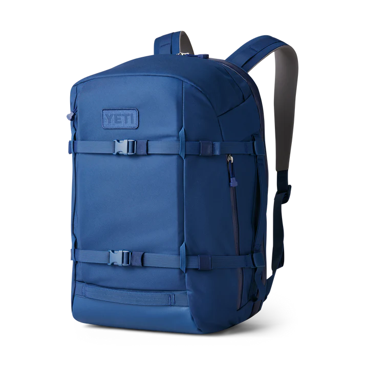 Der YETI® Rucksack Crossroads® 35L - Navy verfügt über gepolsterte Schultergurte, Kompressionsriemen mit Schnallenverschluss an der Vorderseite, eine seitliche Reißverschlusstasche und einen Aufnäher mit geprägtem YETI-Logo - robust für Abenteuer und auf weißem Hintergrund.