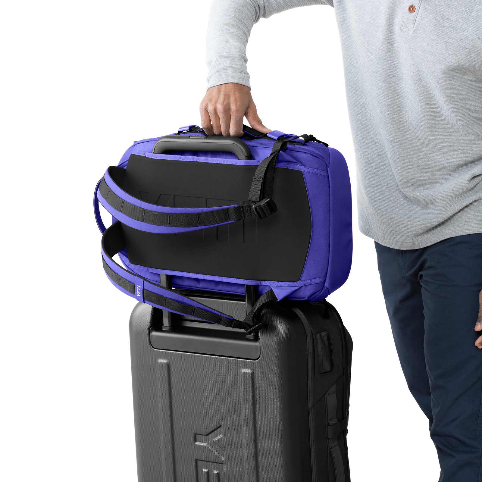 Eine Person in einem grauen langärmeligen Hemd legt einen marineblauen YETI® Rucksack Crossroads® 35L auf einen schwarzen Rollkoffer. Nur ein Teil der Person ist sichtbar, und die Taschen sind gestapelt.
