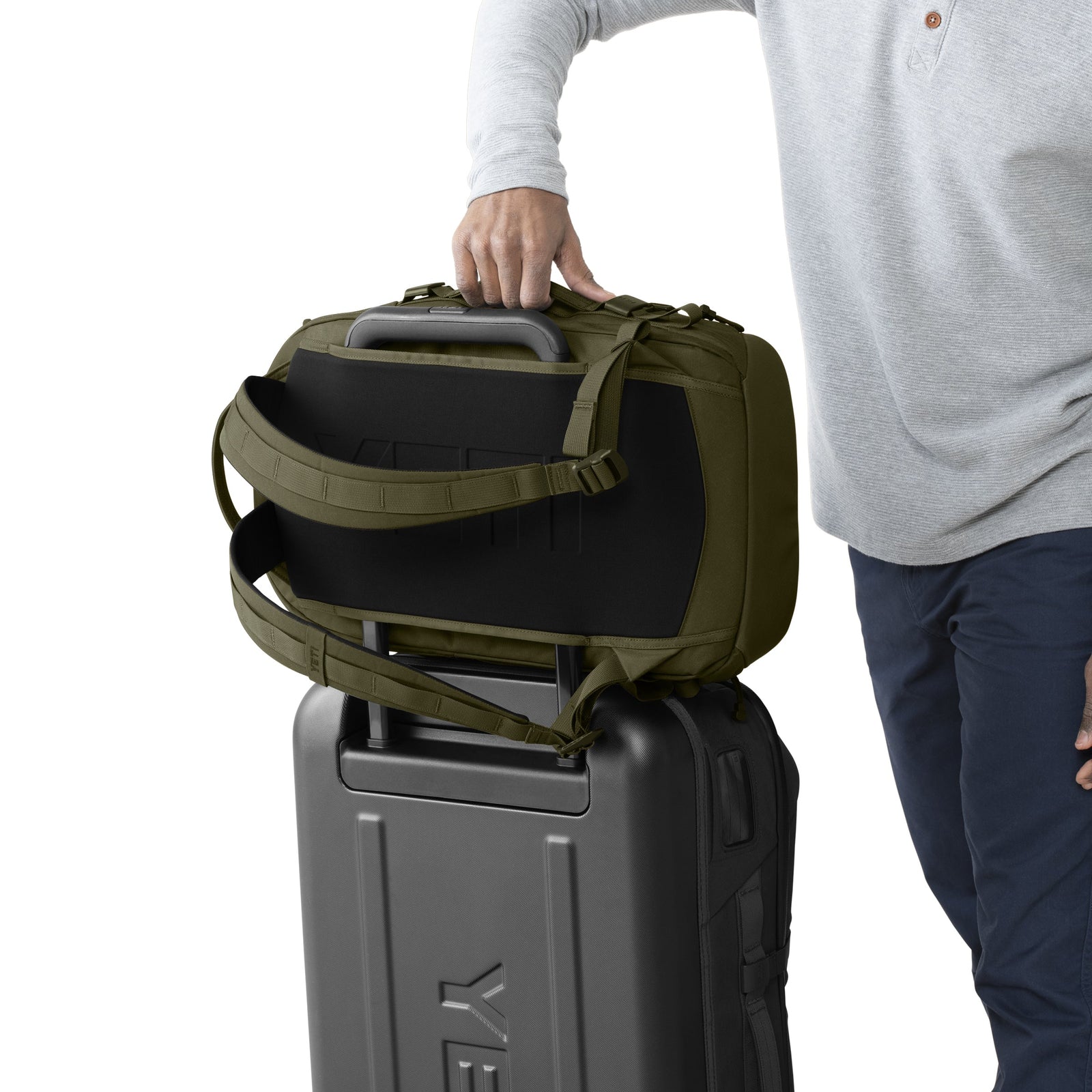 Eine Person, die ein hellgraues Hemd trägt, setzt einen YETI® Rucksack Crossroads® 35L - Olive auf den Griff eines schwarzen Rollkoffers; nur ihr Oberkörper und ihr Arm sind sichtbar.