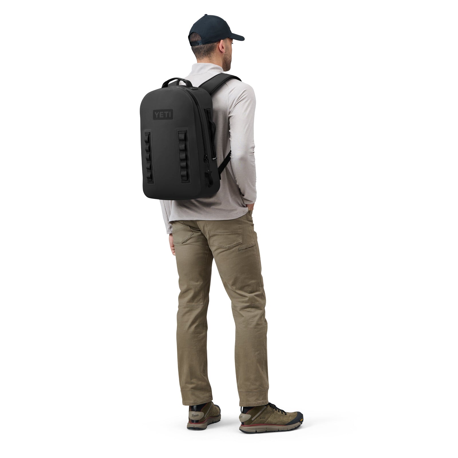 Ein Mann, der von hinten zu sehen ist, trägt einen schwarzen YETI® Rucksack Panga 28, ein hellgraues langärmeliges Hemd, eine olivfarbene Cargohose, Wanderschuhe und eine dunkle Baseballmütze.