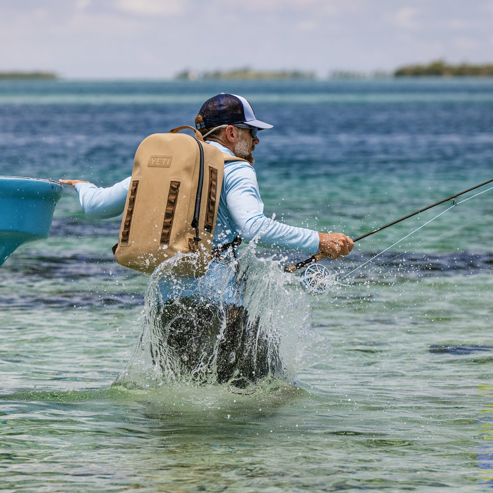 Ein Mann mit Mütze und YETI® Rucksack Panga 28 - Tan watet beim Fliegenfischen durch seichtes, klares blaues Wasser und plantscht in Ufernähe, während im Hintergrund das Land zu sehen ist.