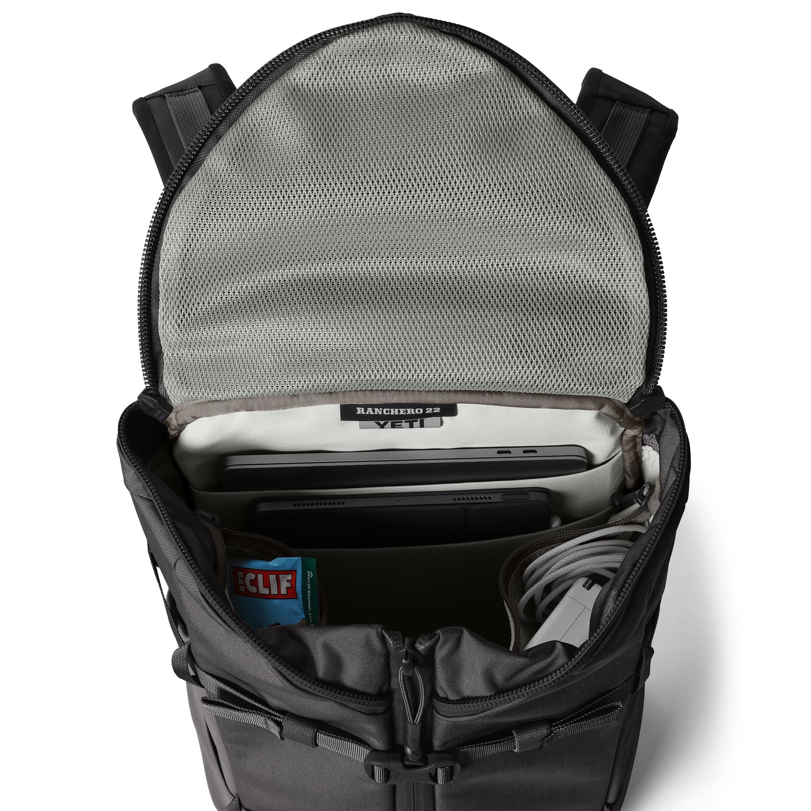 Ein offener schwarzer YETI® Rucksack Ranchero™ 22L bietet Platz für einen Laptop, ein Tablet, ein Notebook, Ladekabel und einen Clif Bar, der in einer Netztasche mit Reißverschluss auf dem Deckel untergebracht ist.