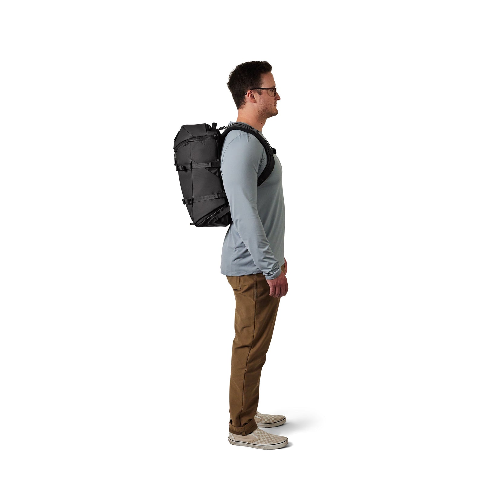 Ein Mann im Profil vor weißem Hintergrund trägt eine Brille, ein hellgraues langärmeliges Hemd, eine braune Hose, weiße Turnschuhe und trägt den YETI® Rucksack Ranchero™ 22L - Black von YETI.