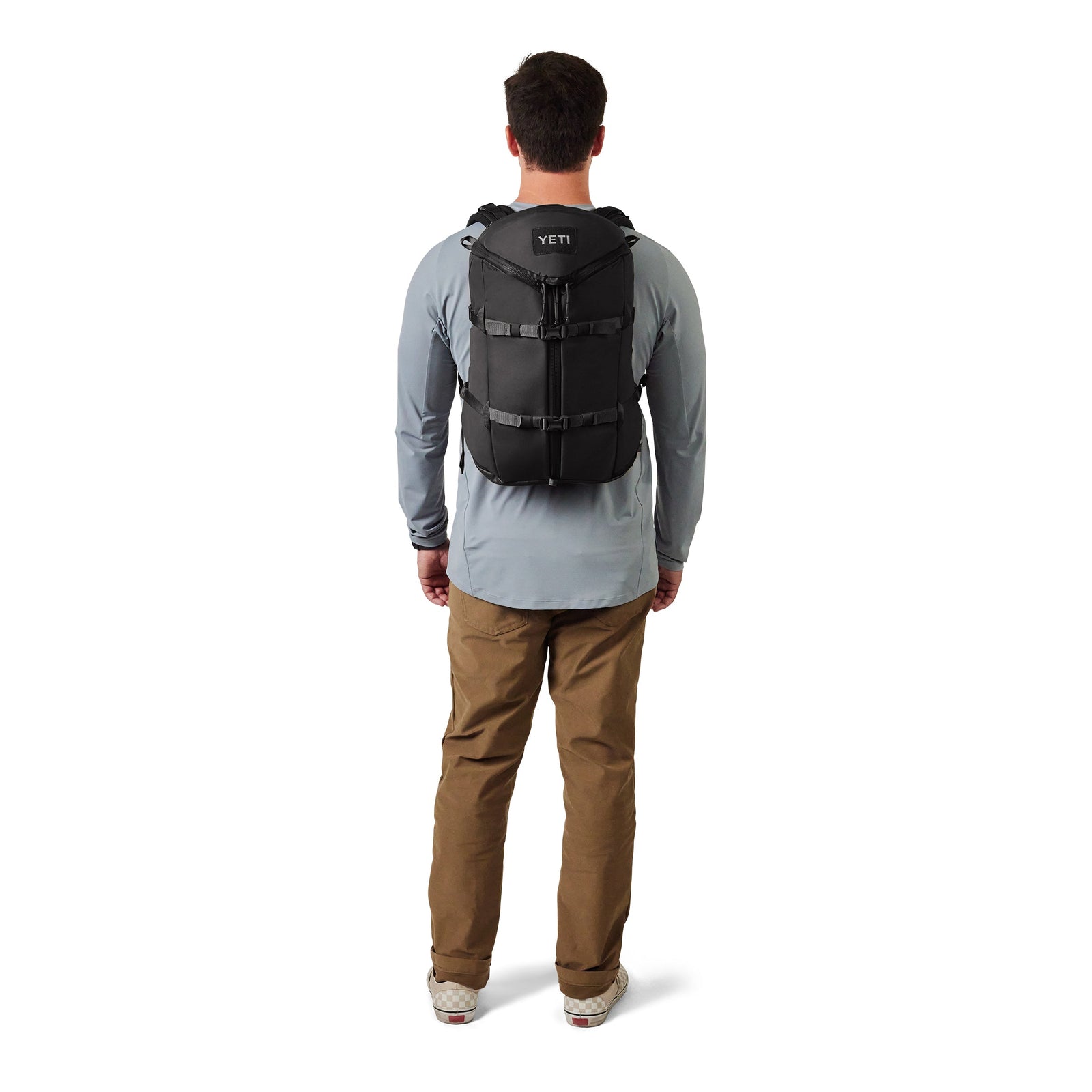 Eine Person in einem hellblauen, langärmeligen Hemd, einer khakifarbenen Hose und hellen Turnschuhen steht mit dem Rücken zugewandt und präsentiert den YETI® Rucksack Ranchero™ 22L - Black auf dem Rücken.