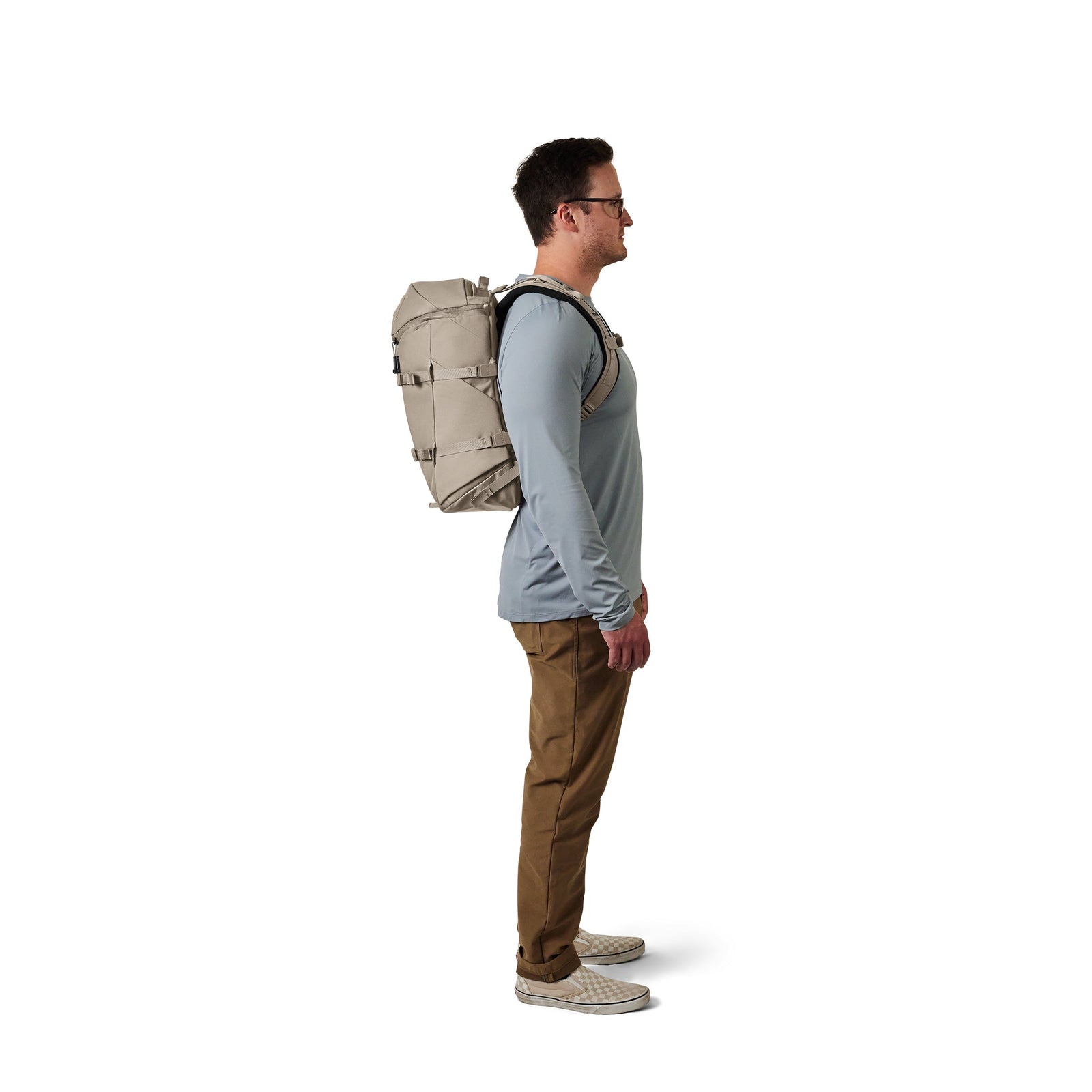Ein Mann im Profil nach rechts trägt eine Brille, ein hellblaues langärmeliges Hemd, eine braune Hose, weiße Turnschuhe und hat einen YETI® Rucksack Ranchero™ 22L in Cape Dark Taupe vor einem schlichten weißen Hintergrund dabei.