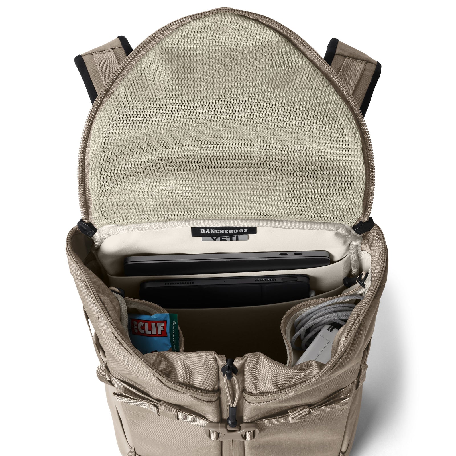 Der YETI® Rucksack Ranchero™ 22L in Cape Dark Taupe ist aufgeklappt und bietet Platz für einen Laptop im dafür vorgesehenen Fach sowie für ein Tablet, einen Clif Bar, ein Ladekabel und Zubehör, das im Inneren gut organisiert ist.
