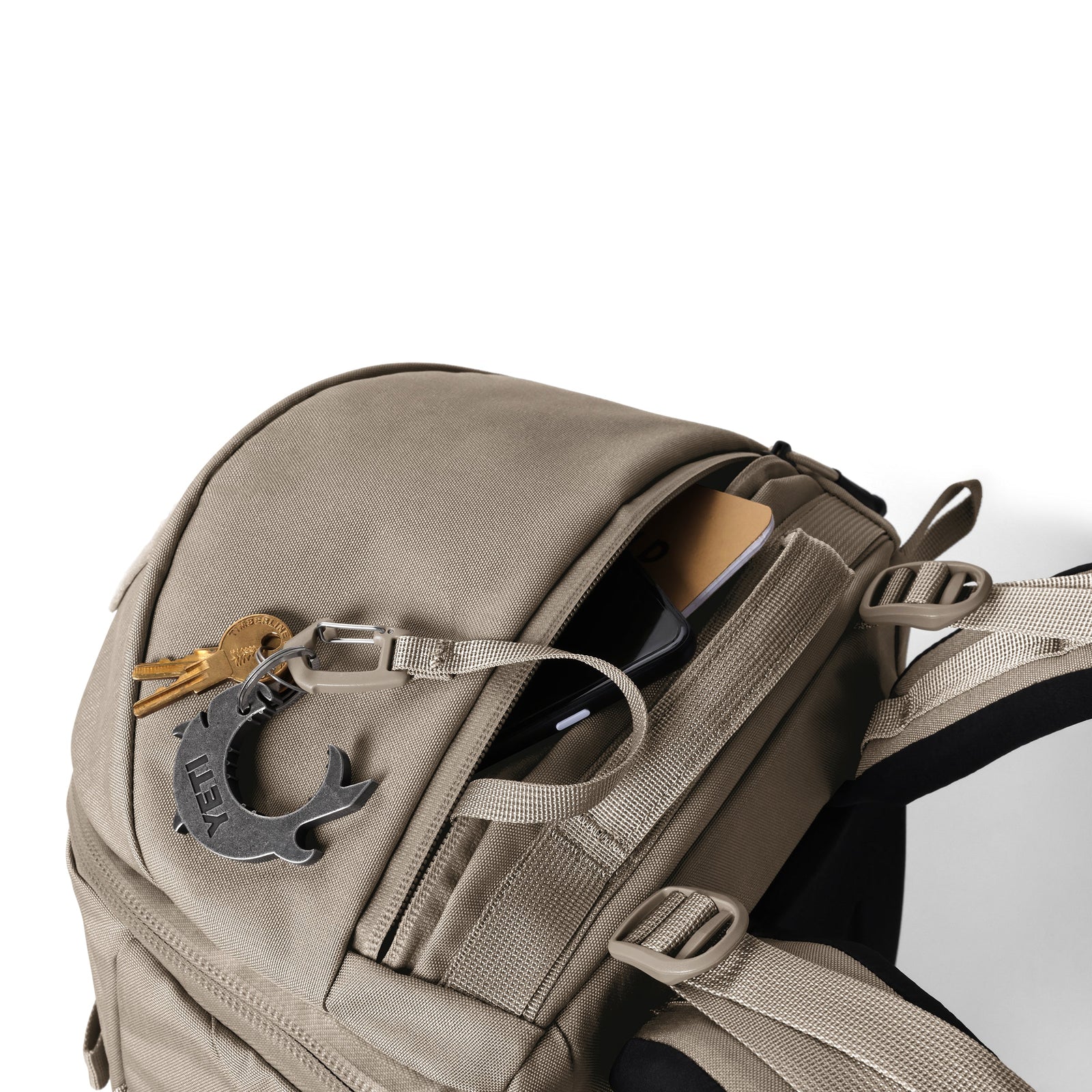 Der YETI® Rucksack Ranchero™ 22L in Cape Dark Taupe verfügt über eine teilweise geöffnete Vordertasche, in der ein Telefon und ein Notebook Platz finden, sowie Schlüssel und ein Multitool aus Metall am Reißverschlusszug. Ein Laptop-Fach sorgt für mehr Komfort und ist auf weißem Hintergrund angebracht.