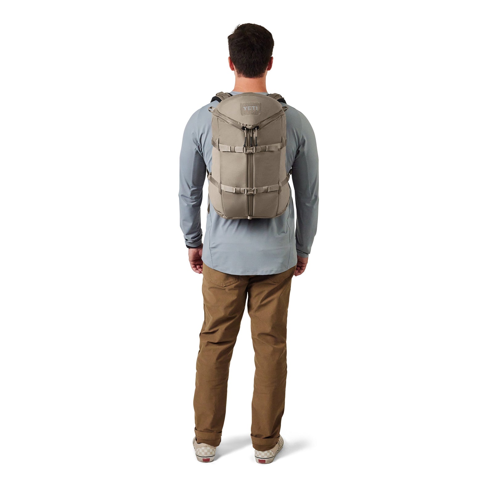 Ein Mann wird von hinten gezeigt, er trägt ein blaues langärmeliges Hemd, eine braune Hose, karierte Turnschuhe und einen YETI® Rucksack Ranchero™ 22L - Cape Dark Taupe von YETI vor einem weißen Hintergrund.
