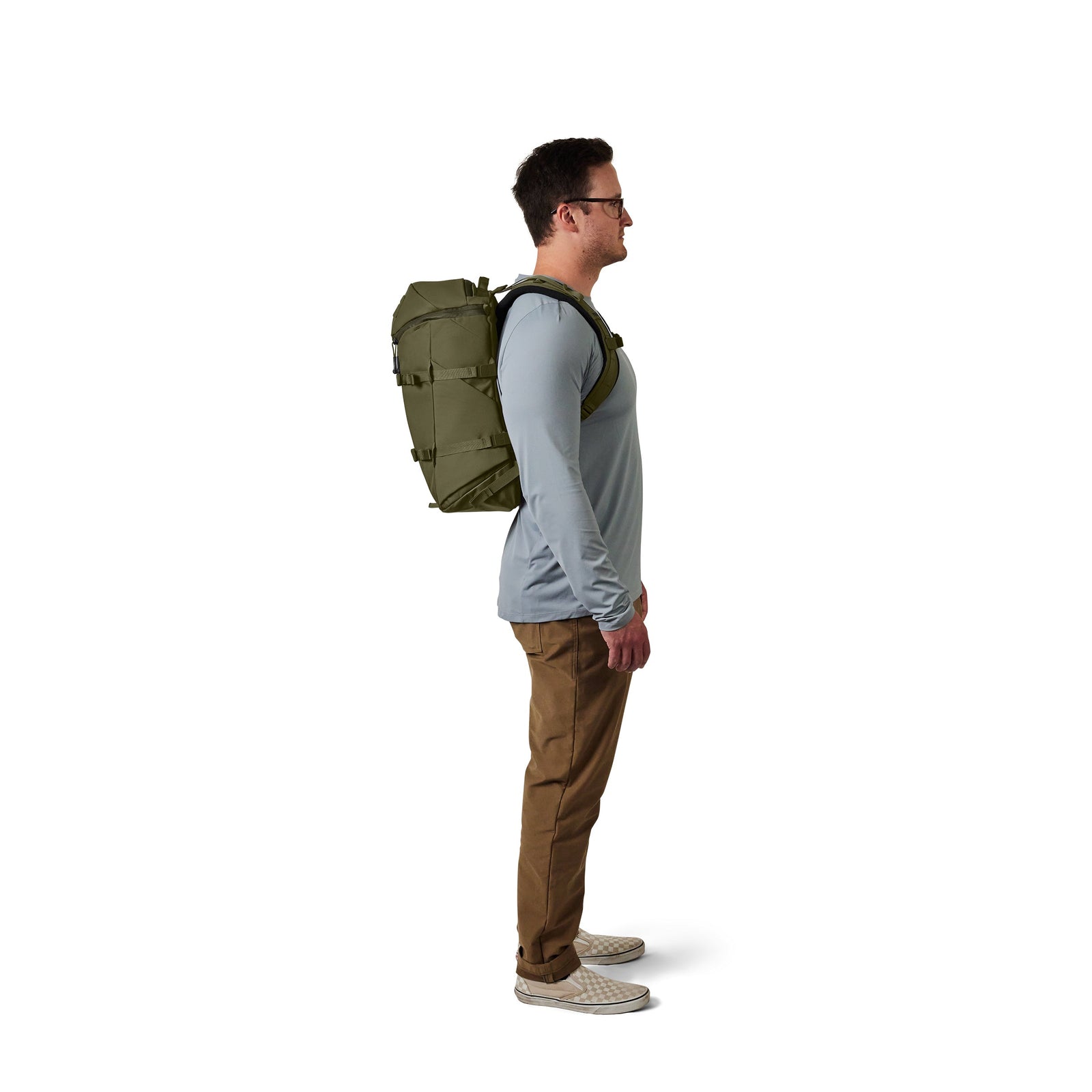 Ein Mann im Profil trägt eine Brille, ein hellblaues langärmeliges Hemd, eine khakifarbene Hose und helle Schuhe. Er trägt den YETI® Rucksack Ranchero™ 22L in Olive mit einem Laptop-Fach aus strapazierfähigem TuffSkin-Nylon vor einem weißen Hintergrund.