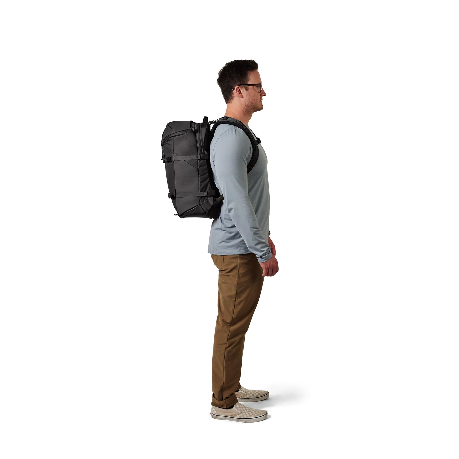 Ein Mann mit Brille, hellblauem langärmeligem Hemd, khakifarbener Hose und Slippern steht im Profil nach rechts und trägt einen schwarzen YETI® Rucksack Ranchero™ 27L von YETI auf dem Rücken vor einem weißen Hintergrund.