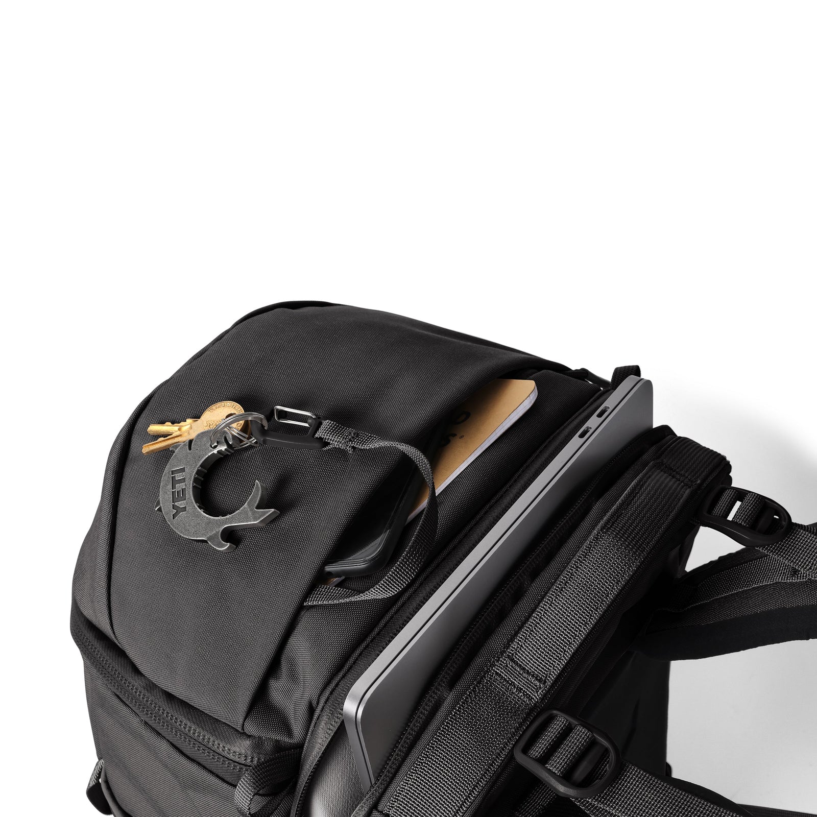 Ein YETI® Rucksack Ranchero™ 27L - Black, teilweise geöffnet, präsentiert im Laptopfach ein Tablet, dazu ein Notizbuch und einen Schlüsselbund mit fischförmigem Anhänger. Der wasserfeste YETI-Rucksack liegt vor weißem Hintergrund.