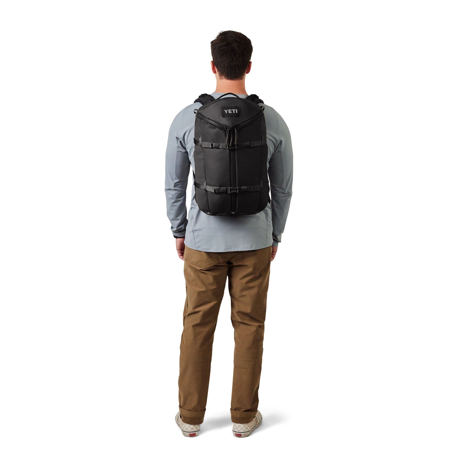 Ein Mann, der ein hellblaues langärmeliges Hemd, eine khakifarbene Hose und Turnschuhe trägt, wird von hinten gezeigt, wie er einen schwarzen YETI® Rucksack Ranchero™ 27L von YETI vor einem schlichten weißen Hintergrund trägt.